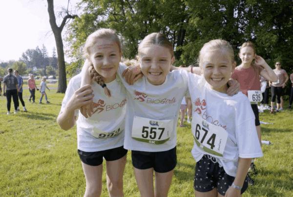 Three smiling girls wearing BIO Girls shirts and race bibs at a 5K event in Fergus Falls